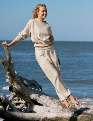 A woman stands barefoot on driftwood at the beach wearing a light beige crewneck sweatshirt and matching sweatpants.