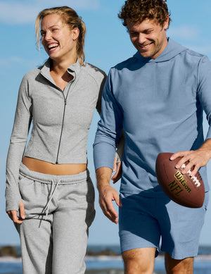 A man and a woman walking outdoors on a beach wearing matching athletic sets. The woman wears a light gray zip-up long-sleeve crop top with matching gray sweatpants.
